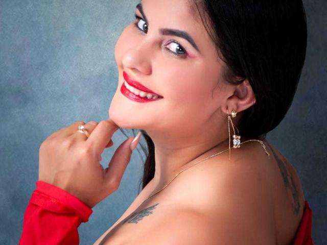 Sharlok with red lipstick, dark hair, earrings, and a red sleeve, showing off her tits with a ring on her finger and tattoos on her shoulder. Smiling, looking back.