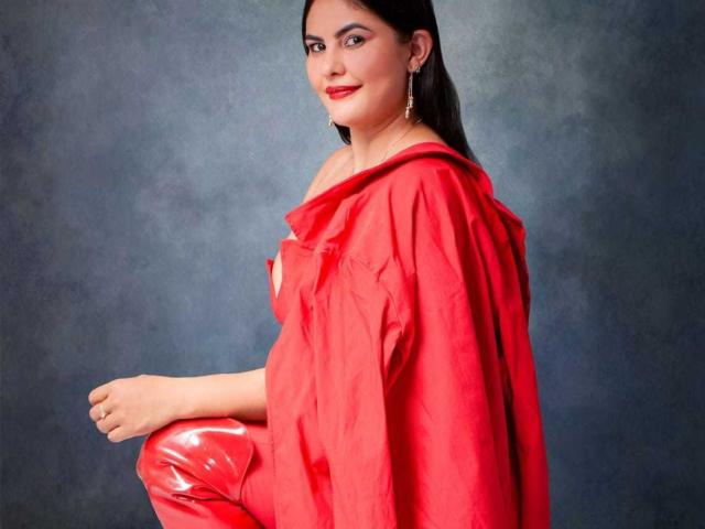 Sharlok's got shiny red latex pants and a loose red shirt, sitting with a cute smile, showing off her ass, with black hair and gold earrings.