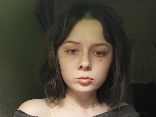 CherryDollye has pale skin, dark wavy hair, small nose, and full pink lips. She's wearing a black off-shoulder top with a silver necklace. Her expression is neutral.