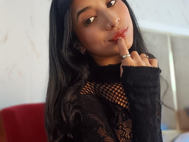 NaiaSanders with long black hair, brown eyes, nose ring, and glossy red lips, wearing a black fishnet top, touching her finger to her lips, showing a ring.