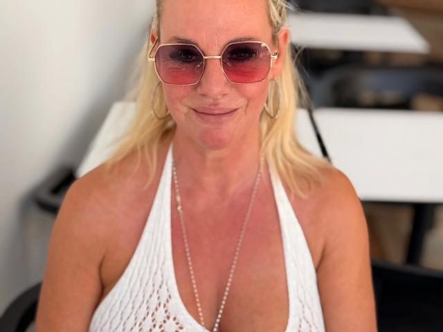 Blonde Blueberry-10-hot with pink-tinted sunglasses, white halter top showing her tits, and a pearl necklace, smiling slightly. Sheer background.