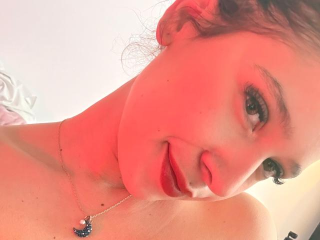 ValeryScot's face with red lighting, showing a slight smile, pink lips, green eyes, and a necklace with a blue and white pendant, bare shoulders visible.