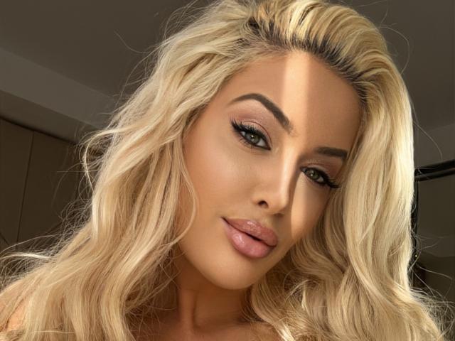 BlancheSummer has blonde wavy hair, full pink lips, and thick black eyebrows. She's looking at the camera with green eyes, long lashes, and a slight smile.