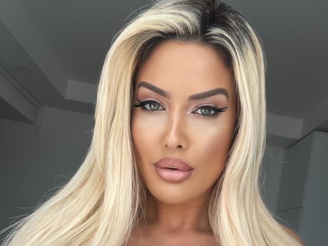 BlancheSummer has blonde hair, green eyes, and full pink lips. She's looking at the camera with dramatic black eyeliner and well-groomed eyebrows. Her skin is tan and glossy.