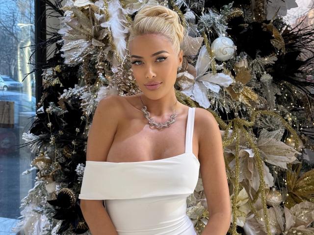 BlancheSummer with blonde updo, wearing off-shoulder white dress, showcasing her tits, standing in front of a glittery Christmas tree, wearing a silver necklace, with a confident smile.