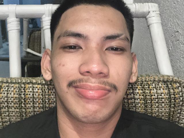 HiddenDesireAru with a light brown face, short black hair, small mustache, and puffed pink lips, sitting on a woven chair with white wicker behind his head.