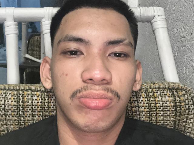 HiddenDesireAru's face with a small mustache, puffy pink lips, and short black hair, sitting in a woven chair with white wooden arms, looking directly at the camera.
