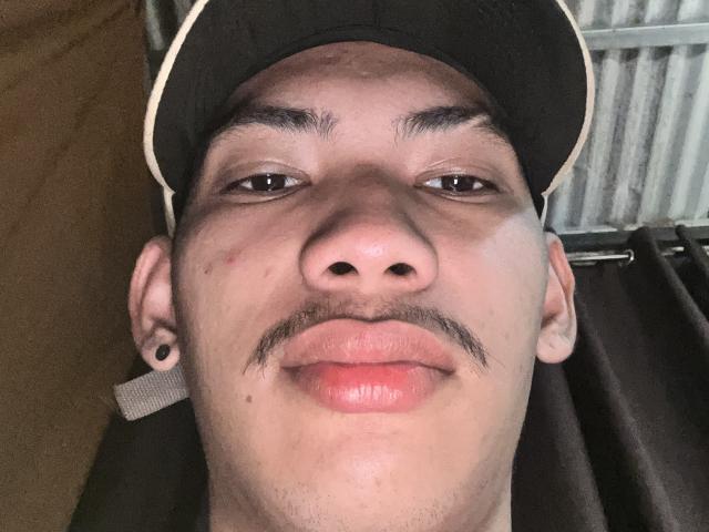 HiddenDesireAru's face with a small mustache, wearing a black baseball cap, ear gauges, and a slight smile, showing a slightly red nose and pink lips.