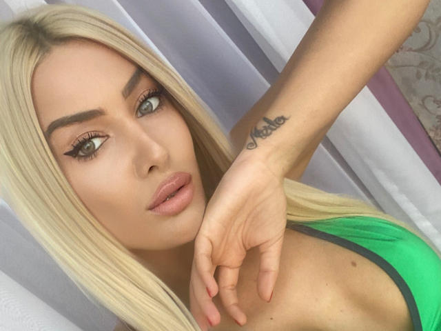 BlancheSummer has blonde hair, green eyes, and a "Kase" tattoo on her right wrist. She's wearing a green top, with her hand near her mouth, showing pink lips.