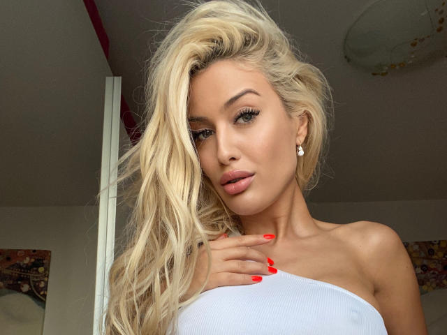 BlancheSummer with long blonde hair, white strapless top, red nails, and pink lips, holding her chest, wearing diamond earrings, has a sexy, attractive, and cute look.