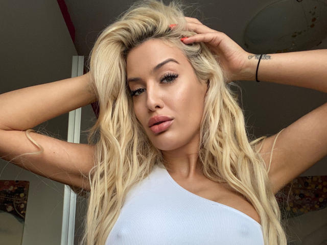 BlancheSummer has long blonde hair, full lips, and wears a white one-shoulder top. She's holding her hair up, showing a tattoo on her right wrist. Her ass is visible.