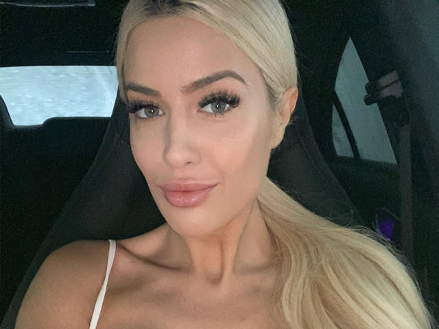 BlancheSummer has blonde hair, full lips, and is wearing a white spaghetti strap top, showing her tits, with long eyelashes and a slight smile in a car.