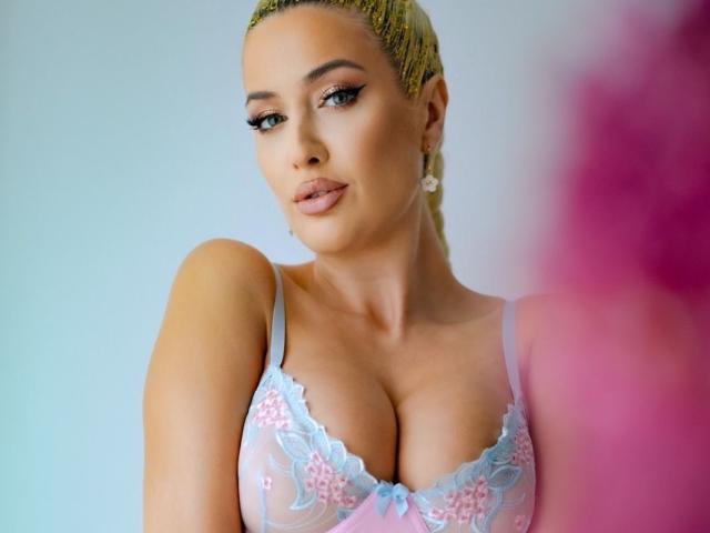 BlancheSummer has blonde braids, large tits in a sheer pink and blue lace bra, and is looking at the camera with a slight smile, wearing dangling earrings.