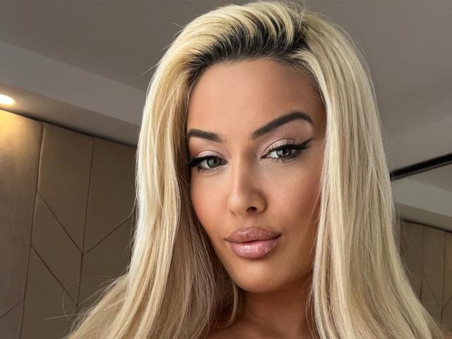BlancheSummer has blonde hair, brown eyebrows, and glossy pink lips. She's looking at the camera with a subtle smile. Her makeup includes long eyelashes and pink eyeshadow.