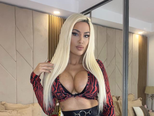 BlancheSummer has long blonde hair, big tits, and a red-black patterned crop top, showing lots of cleavage. She's standing in a room with mirrored walls.