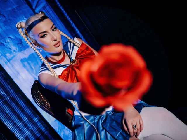 BlancheSummer with braided blonde hair, wearing a sailor outfit with a red bow, extends a blurred red rose towards the camera, sitting on a dark chair, looking sexy.