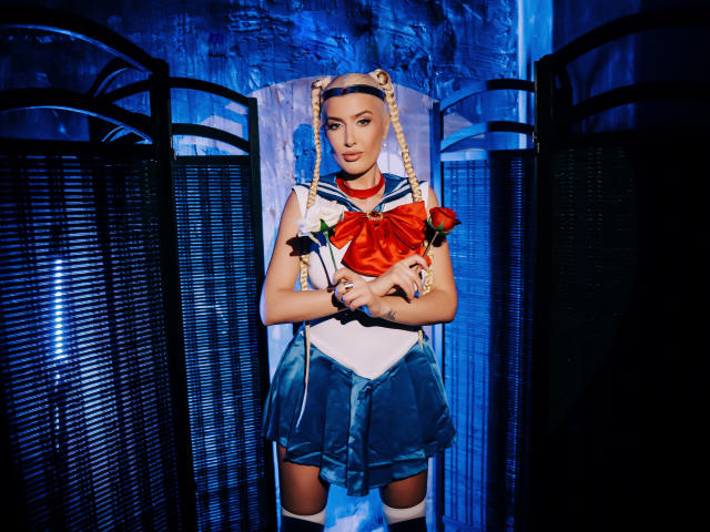 BlancheSummer in a Sailor Moon cosplay, holding red roses, wearing a blue skirt, white top, red bow, thigh-high socks, and braided headband, standing in front of black screens.