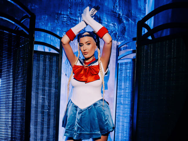 BlancheSummer in a Sailor Moon cosplay, wearing a white top with red bow, blue skirt, white gloves, and red arm bands, standing with arms raised, long braids.