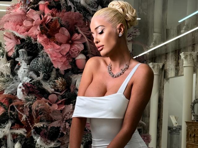 BlancheSummer with blonde bun, wearing white off-shoulder dress, showing tits, necklace, earrings, standing in front of large pink floral arrangement.