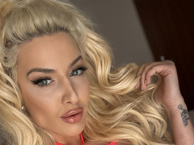 BlancheSummer has blonde curly hair, green eyes, and glossy pink lips. She's holding a lock of her hair, showing a tattoo on her right wrist. Her tits are not visible.