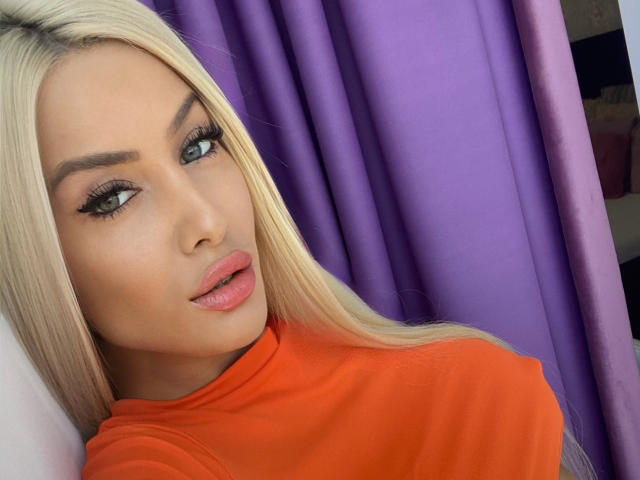 BlancheSummer with blonde hair, green eyes, and full pink lips, wearing an orange shirt, has thick eyelashes and well-groomed eyebrows, looking directly at the camera.