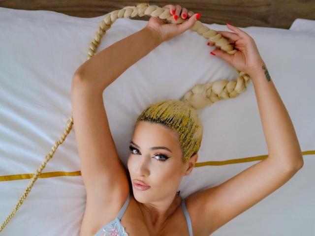 BlancheSummer with long blonde braids, arms raised, lying on a bed, wearing a light blue top, red nails, tattoo on right wrist, looking at the camera.