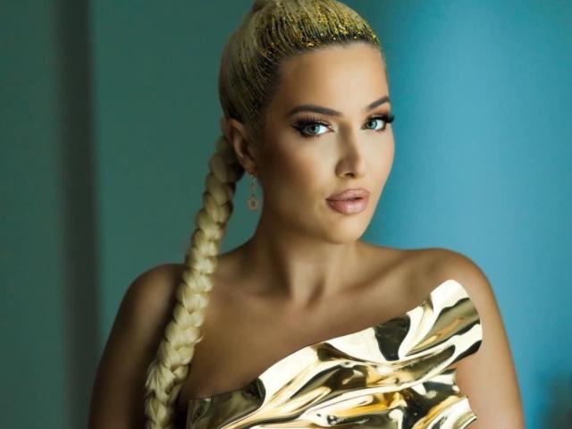 BlancheSummer with blonde braided ponytail, wearing a gold-patterned dress, has tanned skin, full lips, and blue eyes, looking slightly to the side with a neutral expression.