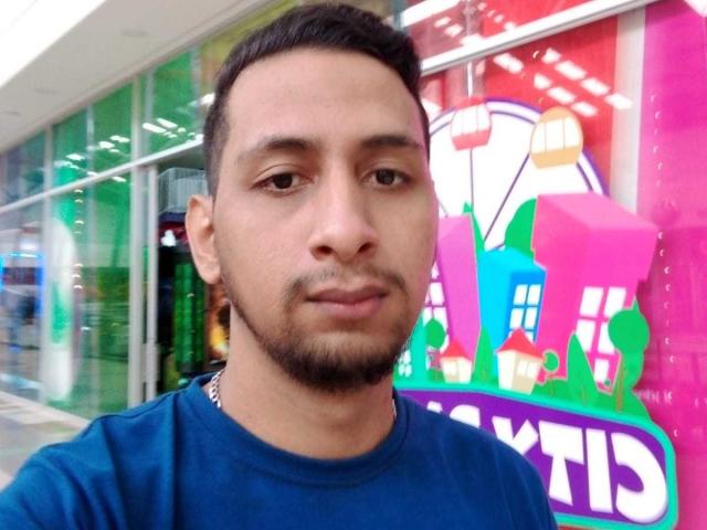 RobinsonSexlv, medium brown skin, short black hair, beard, wearing blue shirt, standing in front of colorful building with balloons, looking slightly to the left, serious expression.