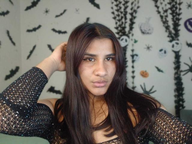 MelinoHottie has long brown hair, wearing a black mesh top, with her hand in her hair, brown eyes, and a neutral expression. Her tits are slightly visible.