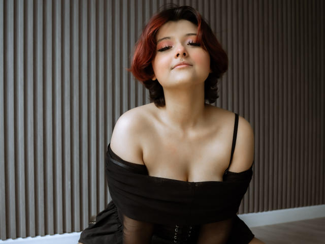 MisakiWong with red hair, cute face, and black dress off-shoulder, showing her tits, sitting with a relaxed, confident smile. Her eyes are half-closed, looking down.