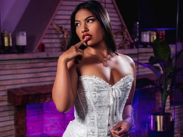 KimJohnsson, con i capelli scuri, la pelle abbronzata e il rossetto rosso, indossa un corpetto bianco senza spalline, con un dito sulle labbra, e appare seducente, con il seno in bella mostra..