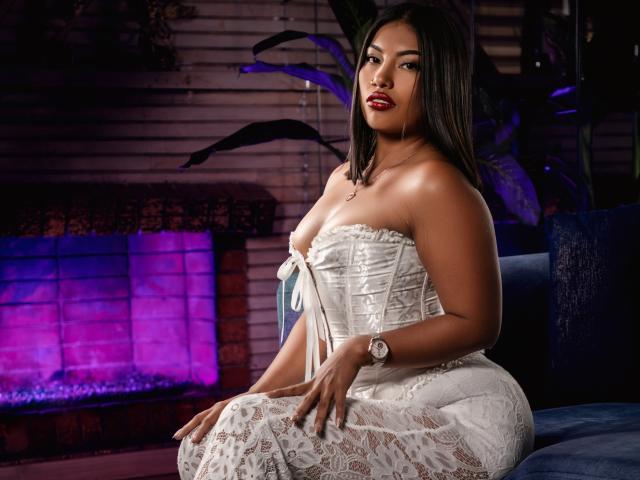 KimJohnsson, una mujer latina de piel marrón media, con cabello lacio y oscuro, vestida con un corpiño y falda blancos de encaje, sentada, mostrando sus senos, con un reloj en la muñeca..