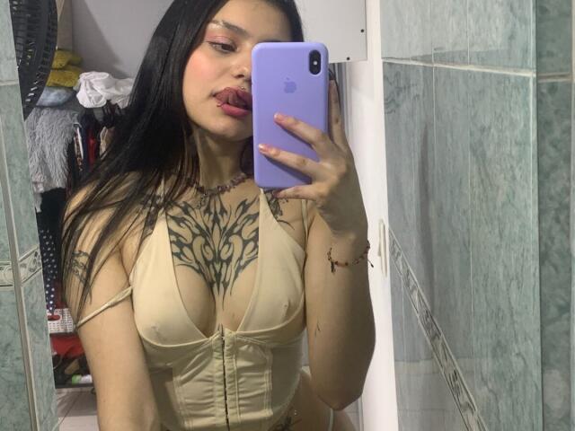 KristyAss with long black hair, pouty red lips, tan top showing her tits, chest tattoo, holding purple phone, mirror selfie, green-tiled bathroom.