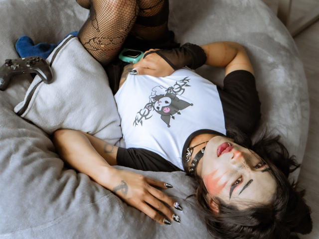 NoemiSky lying on a grey couch, wearing a white and black raglan shirt, black fishnet stockings, and a game controller, with black nails and a black choker.