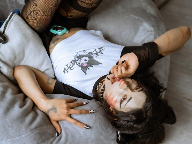 NoemiSky è sdraiata su un letto, indossa una maglietta bianca di cotone, un braccio ricoperto di pizzo nero, calze a rete e una collana. Sta mordendo il dito, mostrando tatuaggi e il viso dipinto di bianco..