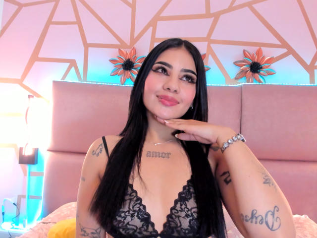 NatallieBlue with long black hair, wearing a black lace lingerie top, smiling, hand on cheek, tattoos on chest and arm, pink headboard, blue lamp, and yellow blanket.