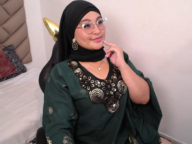 HelueJamil with black hijab, glasses, green dress with gold embroidery, smiling, hand near face, gold earrings, bed with gray and brown pillows in background.