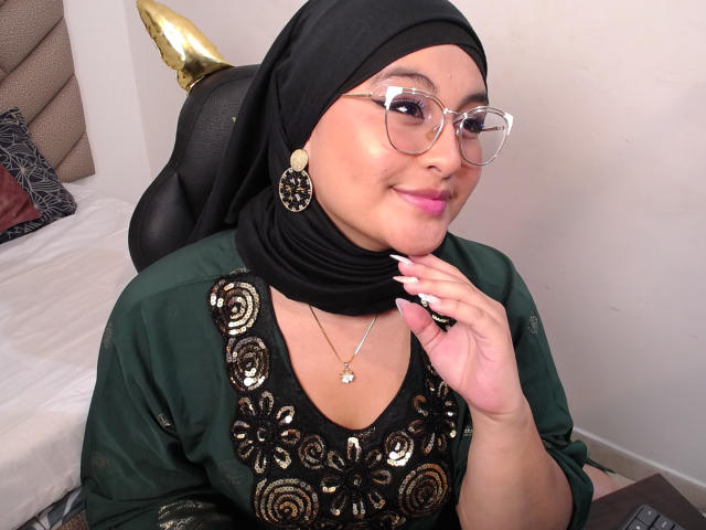 HelueJamil usa um hijab preto, óculos e uma blusa verde escura com bordados dourados, mostrando seus lábios rosados e seu sorriso adorável, com uma mão perto do rosto..