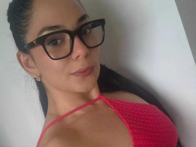 BiancaScott with dark hair in a ponytail, wearing black glasses, pink knit top, showing small tits, subtle smile, gold earrings, and a delicate necklace, against a white wall.