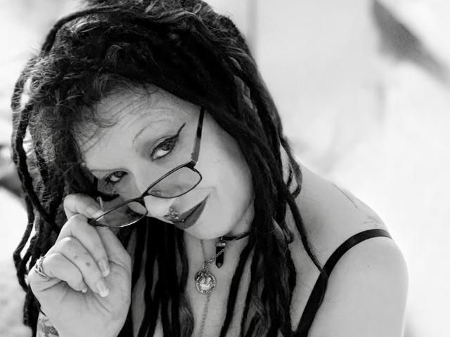 EnchantedLily with dark dreadlocks, wearing glasses, holding them with her right hand, has bold eyeliner, nose ring, and pendant, looking seductively at the camera. Black-and-white photo.