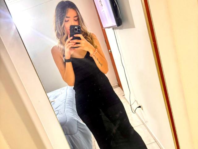 AdharaCooperr takes a mirror selfie, wearing a black sleeveless dress, holding a phone, with wavy blonde hair, showing her tits and ass in the reflection.