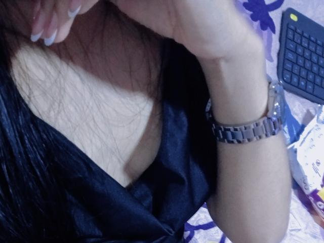 AnaisHilton's tits are visible in a black, low-cut top. She's wearing a silver watch on her right wrist, with her hand near her face. Her long brown hair is visible.