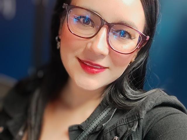 MichellMonet with red lipstick, black hair, and pink glasses, smiling at the camera. She's wearing a black shirt, showing her tits slightly through the open collar.