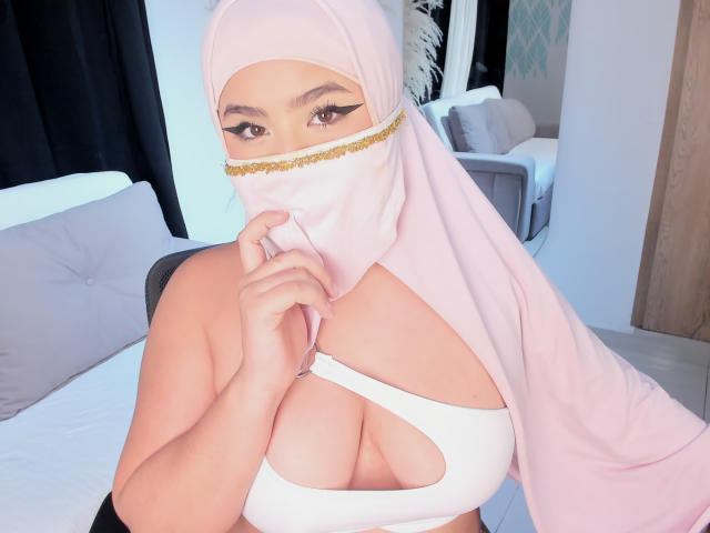 DaliilaAli with pink hijab, white mask, and white cutout top, showing large tits, holding mask, winged eyeliner, soft light, wooden door in background, bedroom setting.