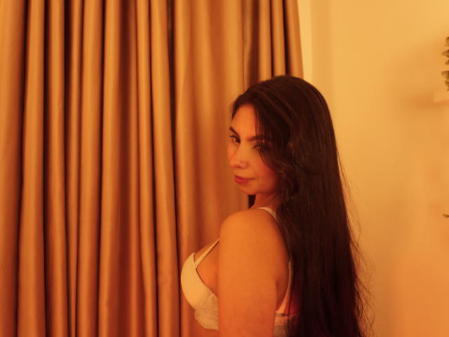 MilaDor, con cabello largo y oscuro, mirando por encima del hombro, vistiendo un sostén blanco, mostrando sus pechos, en una habitación con luz cálida y cortinas beige..
