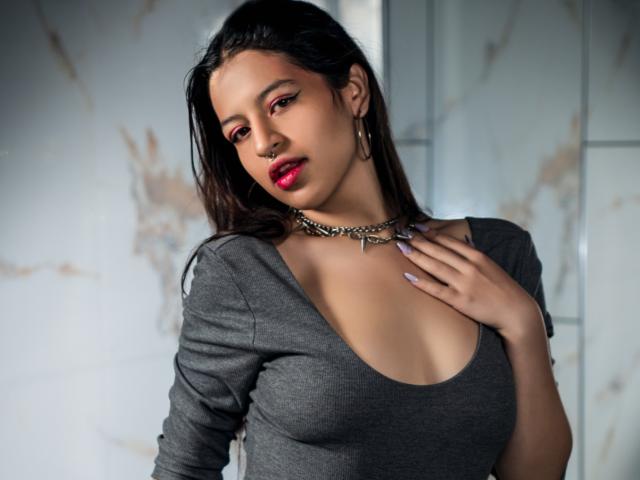DalilaVolvok with dark hair, red lipstick, grey top, showing her tits, silver chain necklace, hand on chest, grey background, marble texture, looking at camera, slightly tilted.