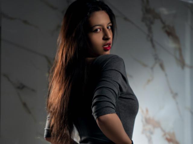 DalilaVolvok with long brown hair, red lips, and a gray shirt, looking over her shoulder, her ass slightly turned to the camera. She's sexy and confident.