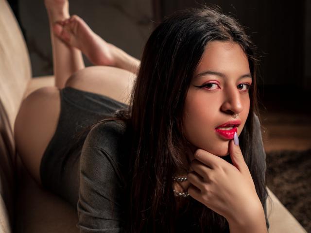 DalilaVolvok lying on a couch, face down, finger in mouth, looking at camera, black dress, red lipstick, nose ring, long dark hair, bare ass, legs up, soft lighting.