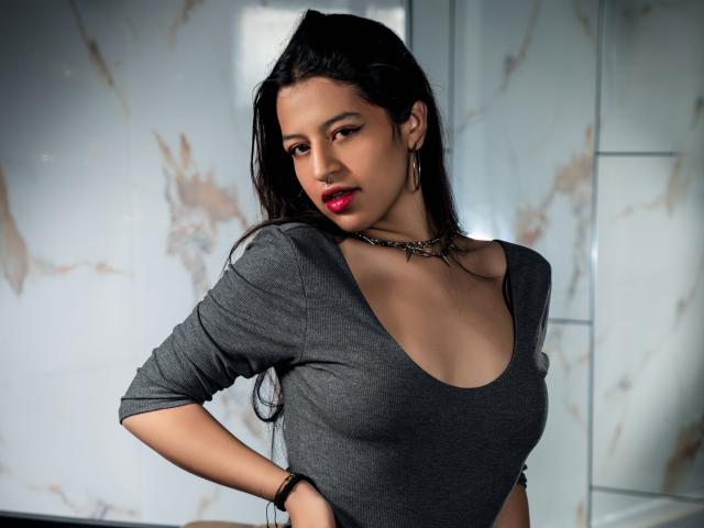 DalilaVolvok with dark hair, red lipstick, gray shirt, chain necklace, and bracelet, showing her tits, hands on hips, looking down, in a modern, light-filled room.