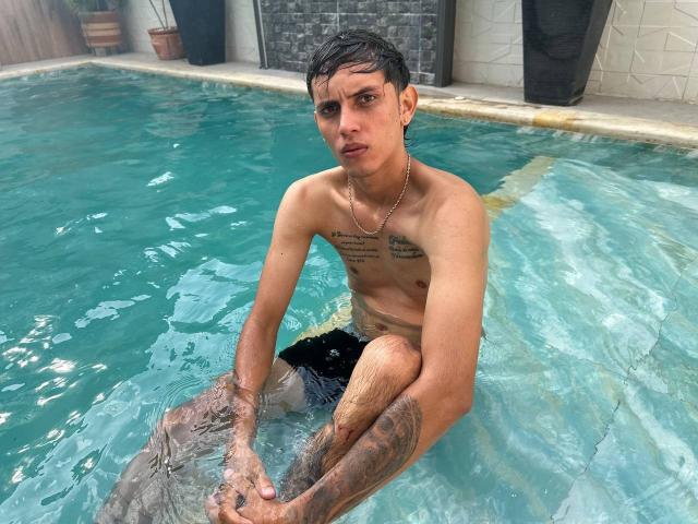DannaXFabian with wet black hair, tattoos on chest and arms, wearing a gold chain, sitting in a blue-tiled pool, looking at the camera with a serious expression.
